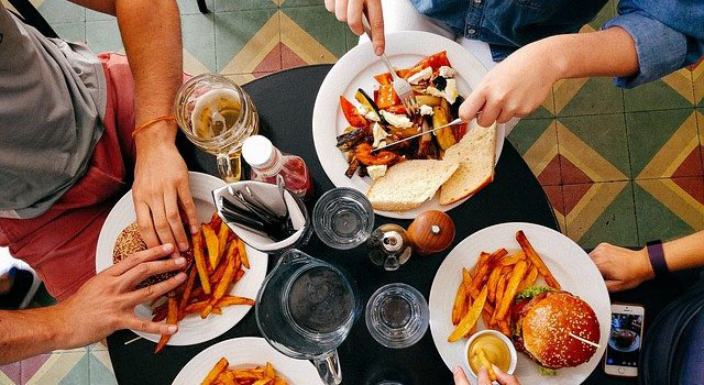 Four people at a restaurant table, three eating burgers and fries, one eating roasted vegetables and bread.