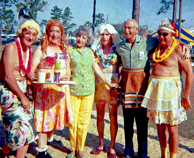 A half dozen senior citizens, five men and a woman, pose at an Airstream park. The woman at center left wears brightly colored pants and blouse. The men are partially or entirely in women's clothing, and one is balancing three cans on a tray which also holds a drinking cup. Behind them are Airstream trailers, pine trees, and a yellow and blue canopy.