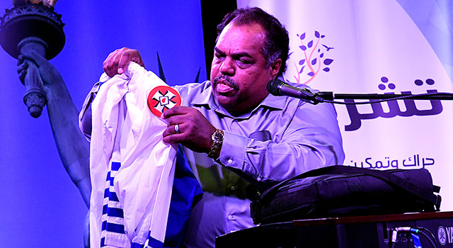 A man seated on a stage, wearing a blue button-down shirt, holds up a white robe adorned with emblems of the Ku Klux Klan