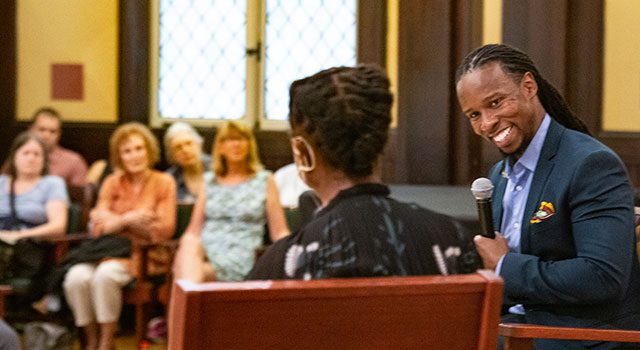 Professor Ibram X. Kendi sits with a microphone, smiling and looking at an unidentified speaker (seen from behind) in a church with a small audience in the background.